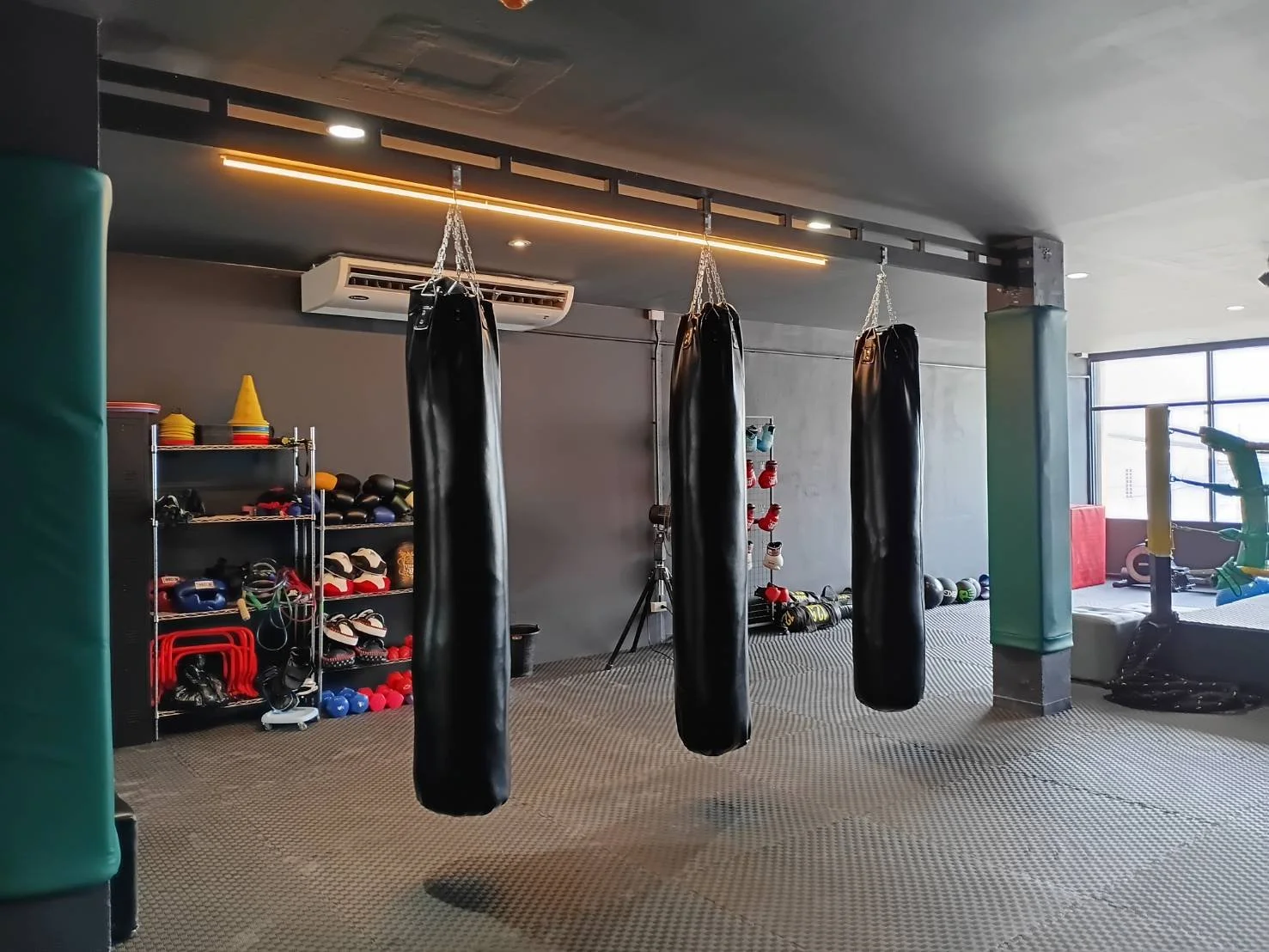 Gym with three punching bags hanging from the ceiling, workout gear on shelves, and equipment on the floor near large windows.