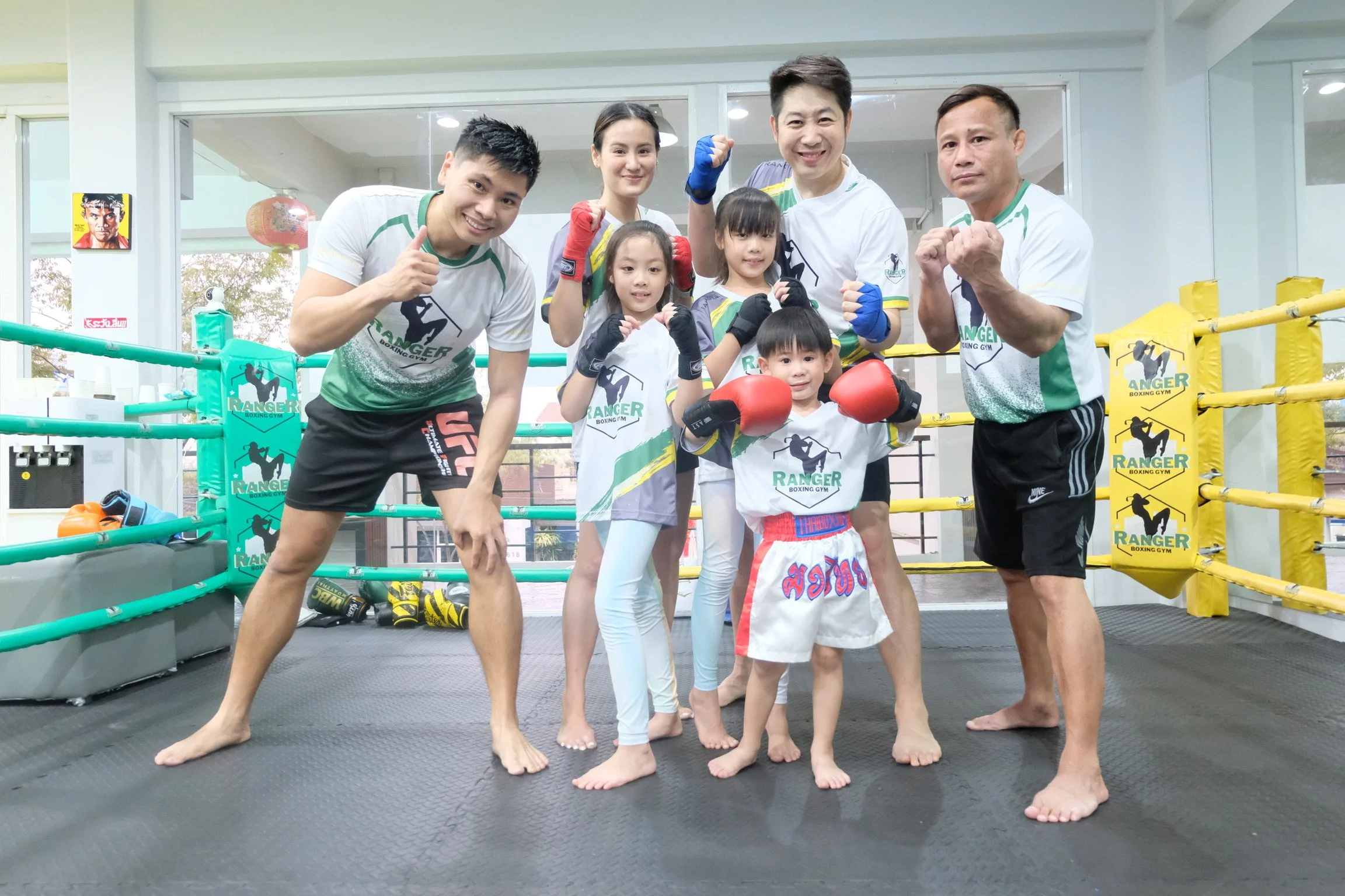 Group of six people in a boxing gym, including children and adults, posing with fists up for a photo. They are dressed in boxing gear and t-shirts with a boxing gym logo. The gym has boxing rings, gloves, and equipment visible in the background.