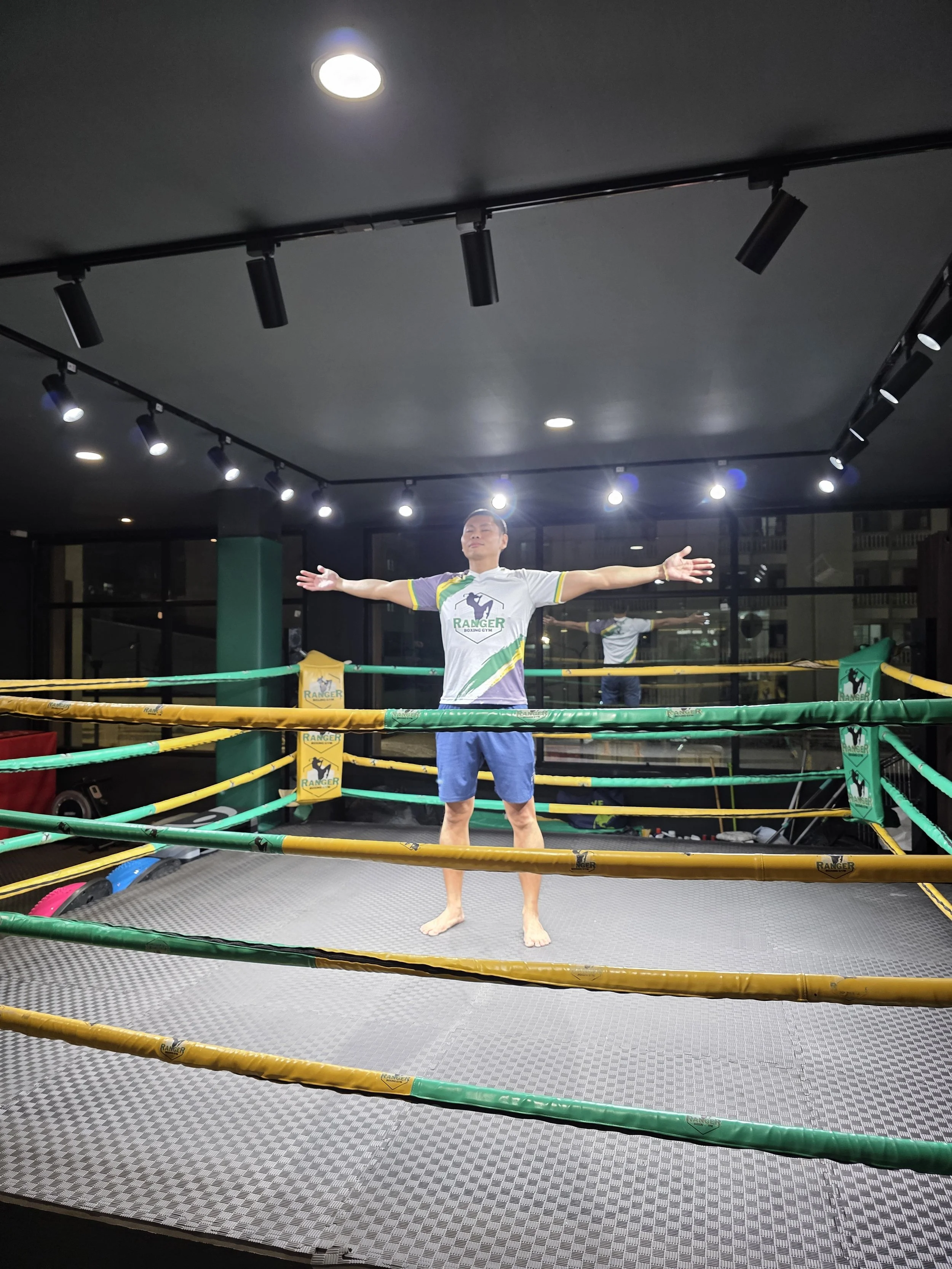 A man stands barefoot inside a boxing ring with arms outstretched, wearing a white and green Ranger boxing gym shirt and blue shorts, with a black floor and overhead lighting.