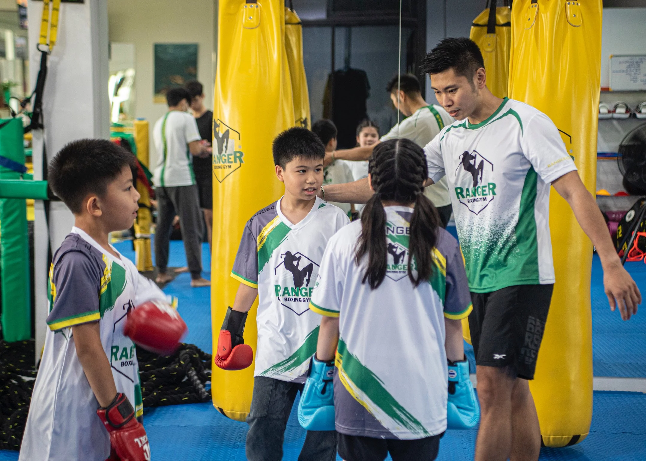 Young boxing students listening to their coach in a boxing gym, wearing matching uniforms with the words 'Ranger Boxing Gym' and a logo of a boxer.