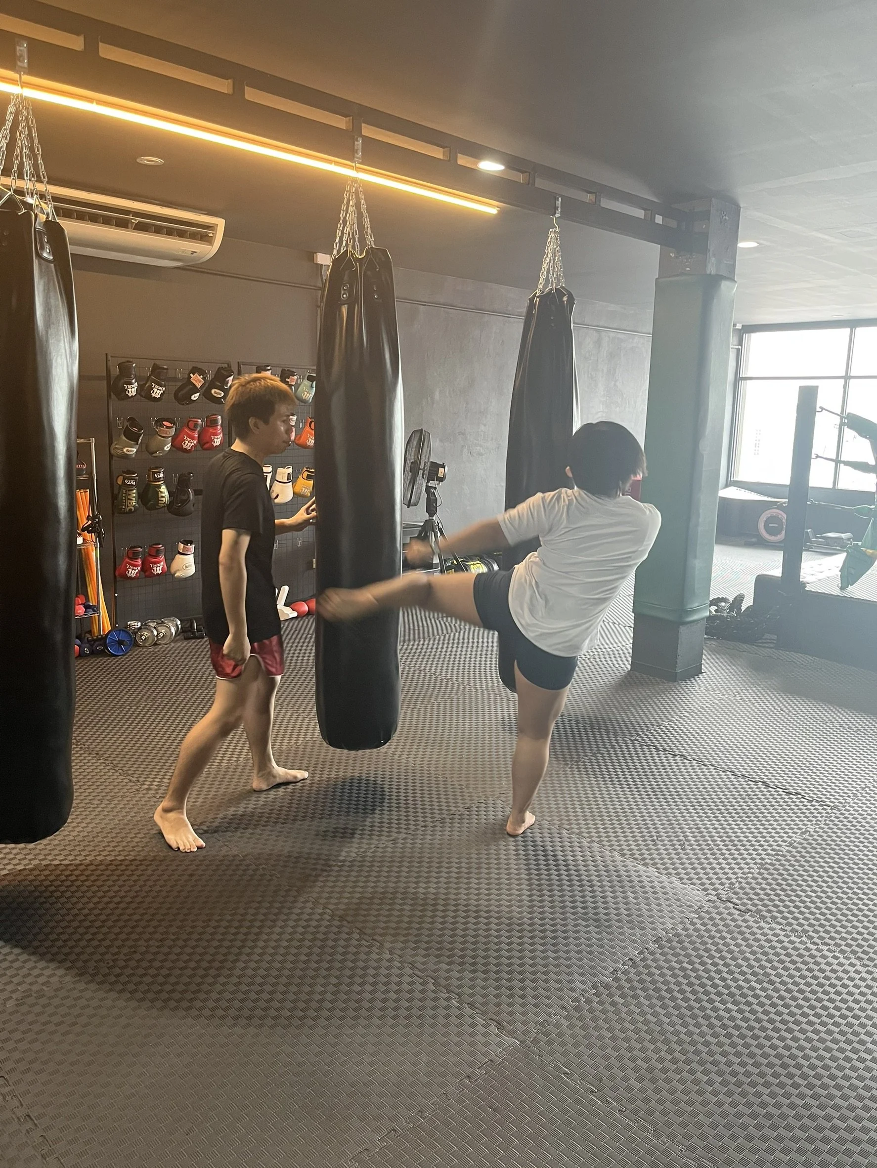 Two men practicing martial arts in a gym, one kicking a punching bag while the other observes. The gym has boxing gloves and other workout equipment on display.