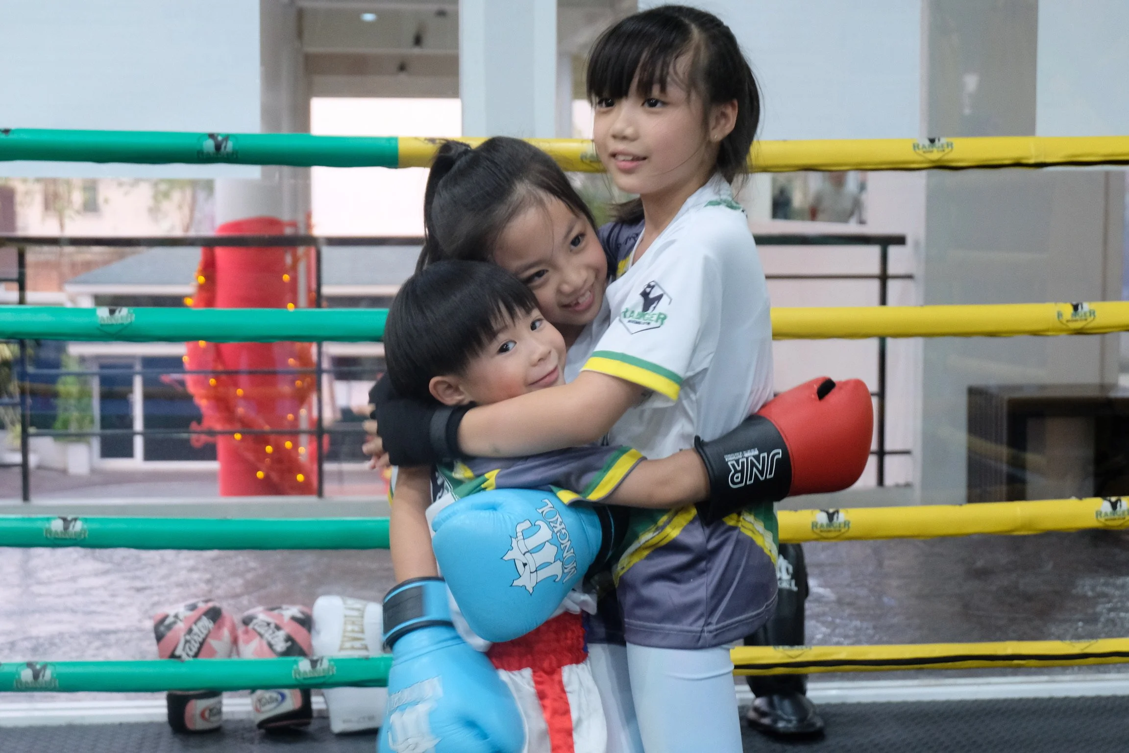 Three young children are hugging each other in a boxing ring. The boy in boxing gloves is hugging two girls, all smiling, with yellow and green ropes around the ring. In the background, there is a red punching bag and other equipment.