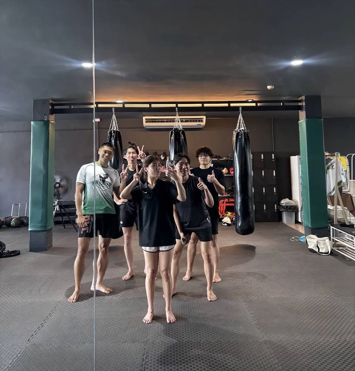 Group of five people in a gym, standing barefoot on a mat in front of punching bags, posing for a mirror selfie with peace signs and thumbs up.