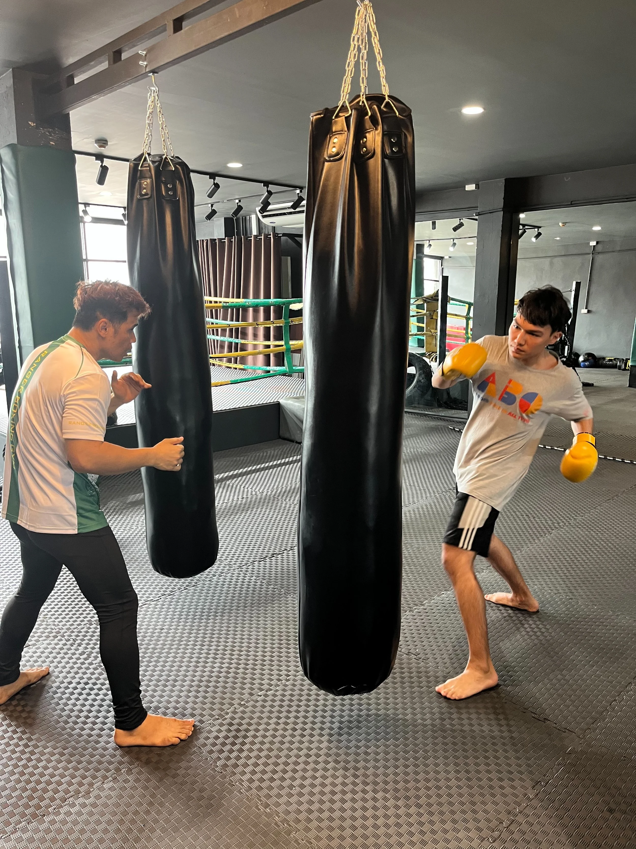 Two men practicing boxing with a black punching bag hanging from the ceiling inside a gym. One man is wearing a white t-shirt with yellow boxing gloves, the other is barefoot, wearing a white t-shirt and black shorts with white stripes, and is in a p