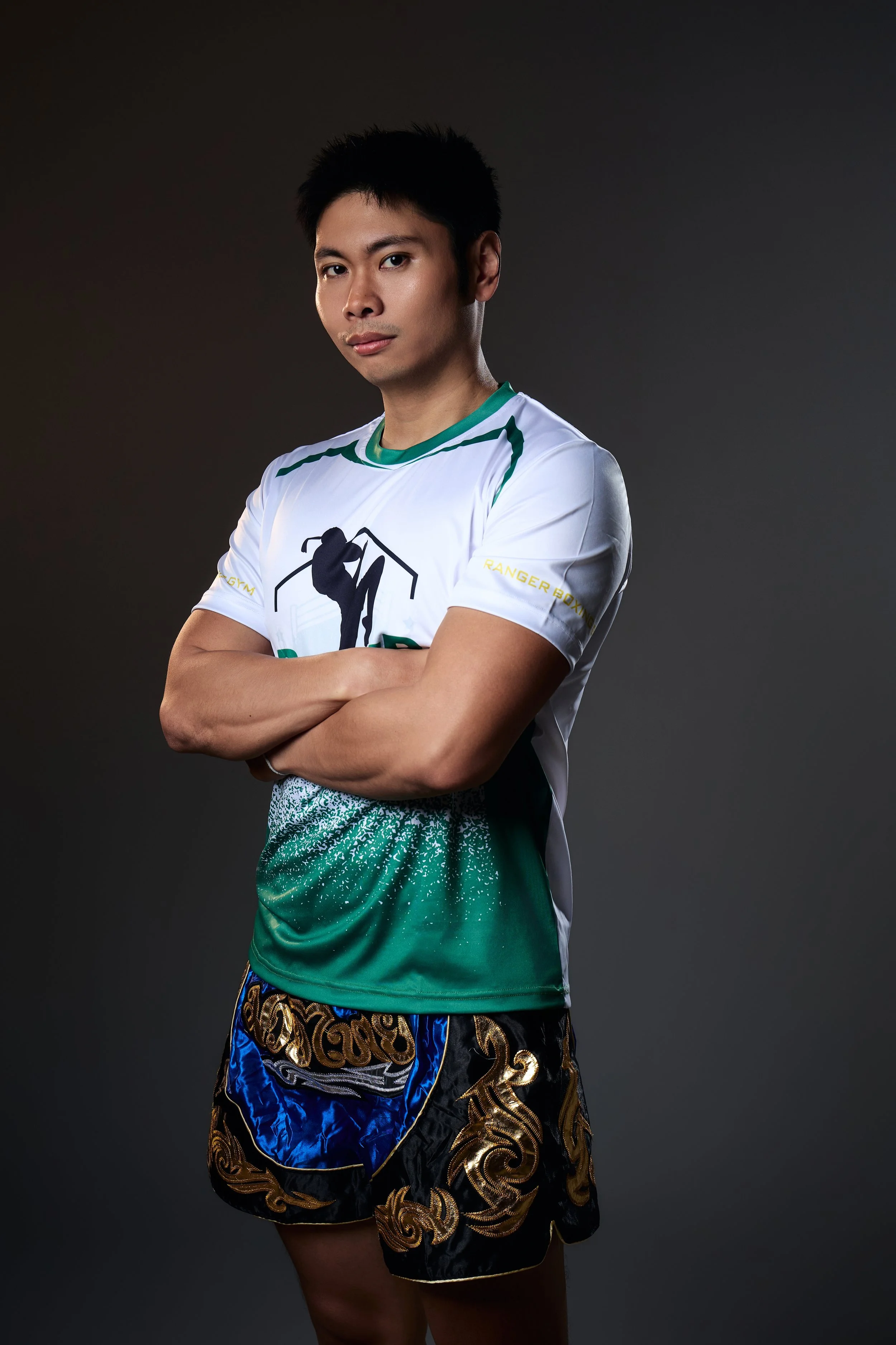 Young man with short black hair wearing a Ranger Boxing Gym shirt and Muay Thai shorts, standing with arms crossed against a dark background.