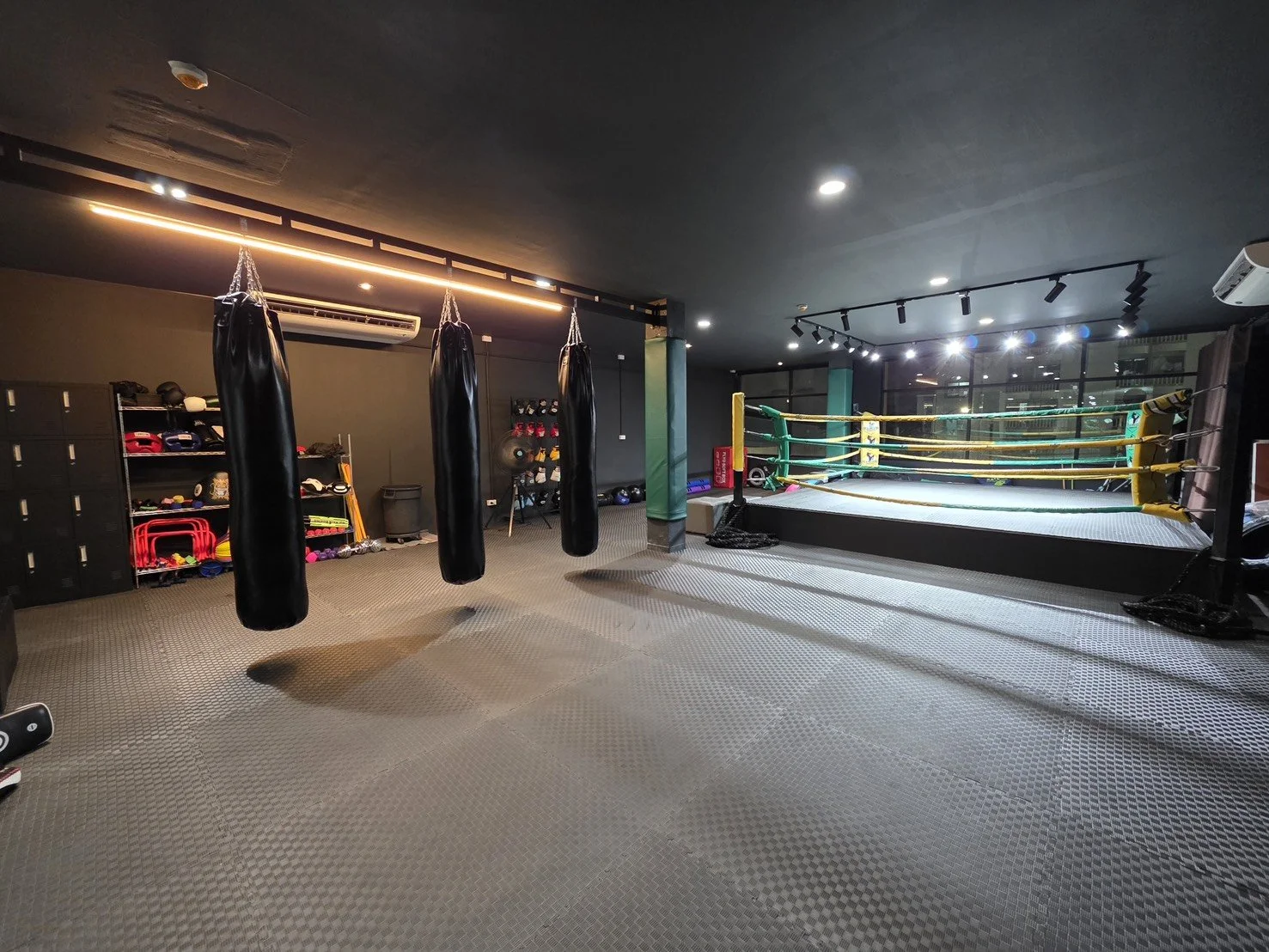 An indoor boxing gym with hanging punching bags, a boxing ring with yellow ropes, and varied equipment such as gloves, helmets, and weights, with dark walls and large windows.