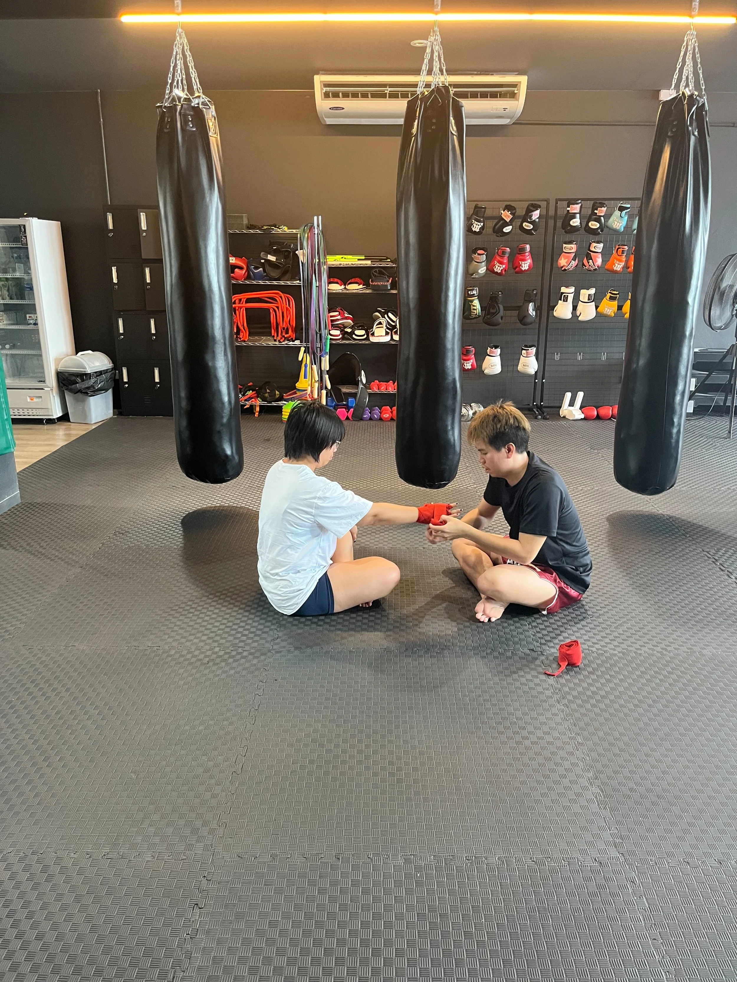 Two people sitting on the floor of a martial arts gym, wrapping each other's hands with red hand wraps. The gym has three black punching bags hanging from the ceiling, and shelves with boxing gloves, headgear, and other training equipment in the back
