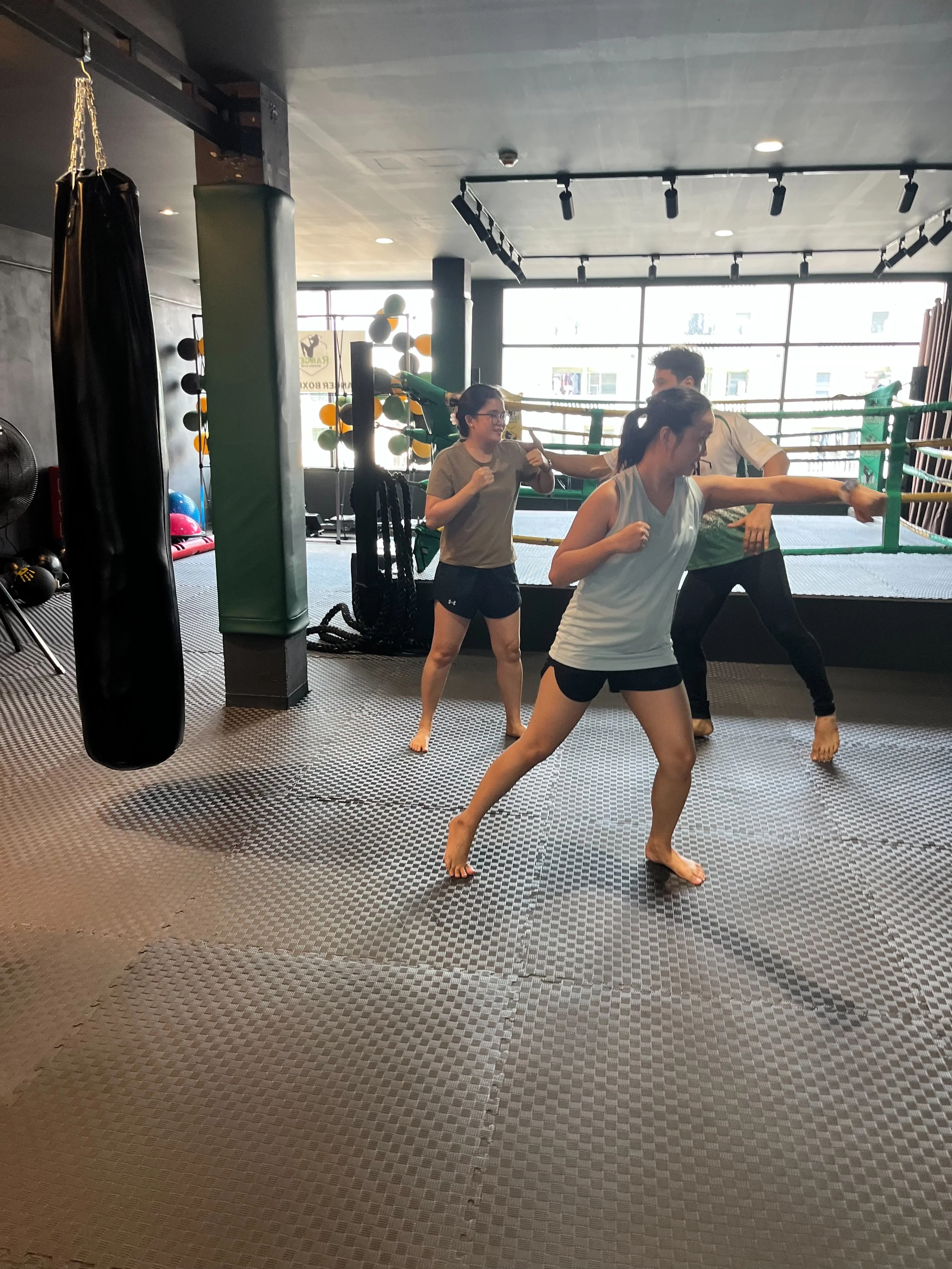 Three women practicing boxing in a gym with a punching bag and a boxing ring.