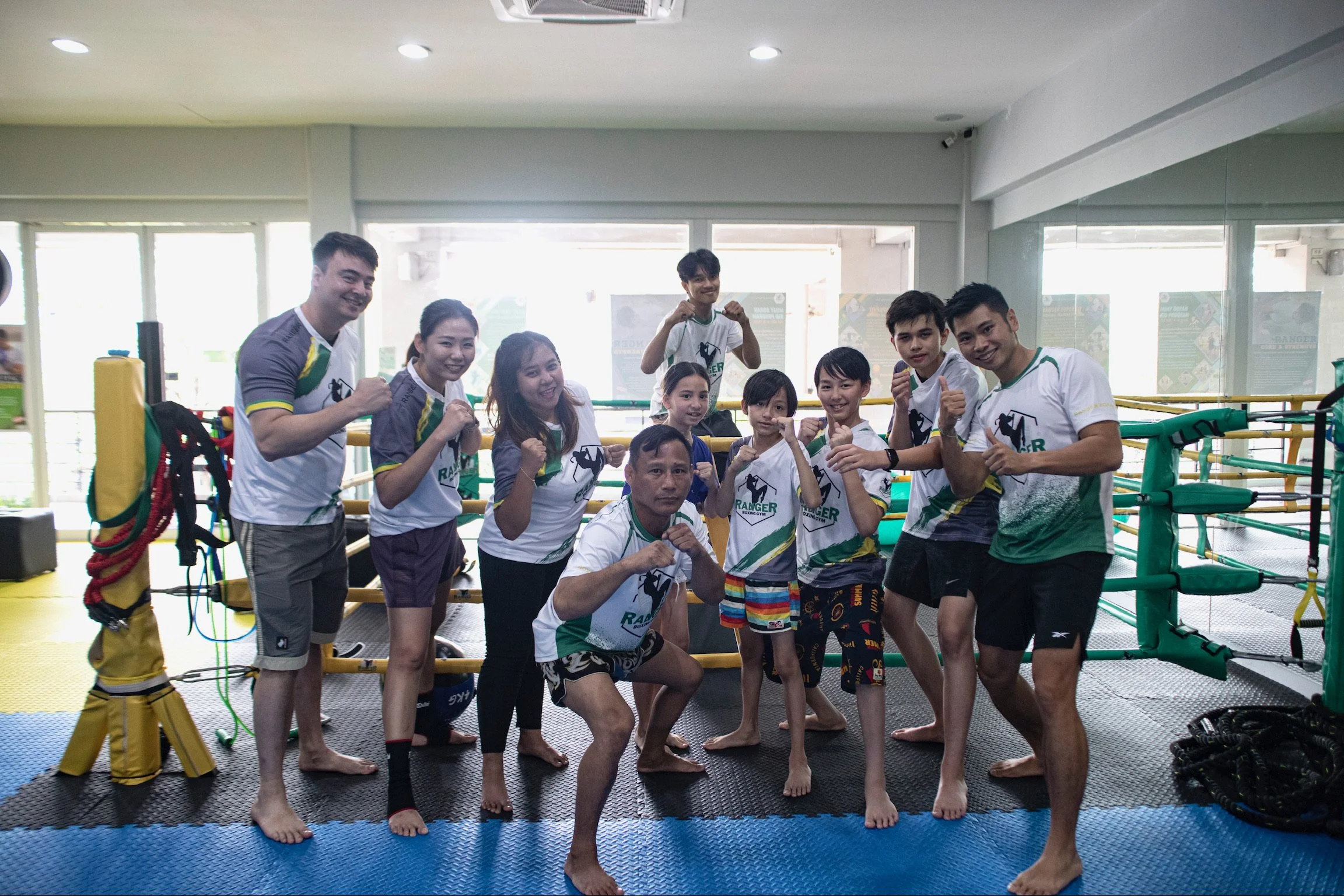 Group of martial artists and fighters inside a gym with boxing ring, some making fists and smiling at the camera.