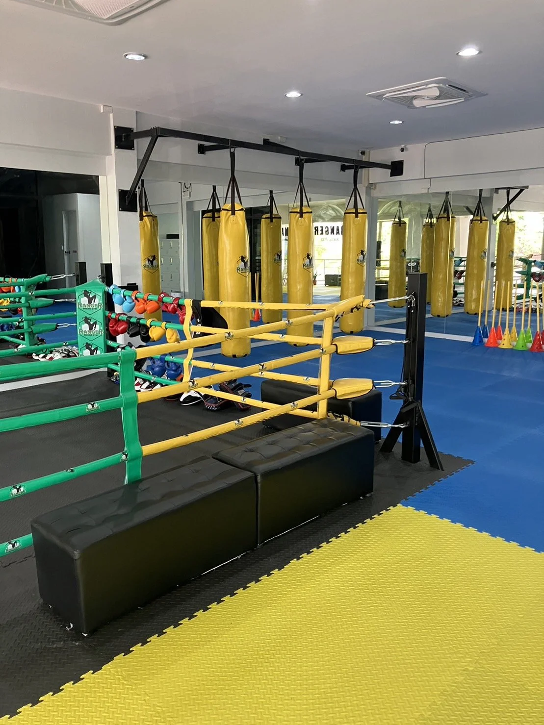 A boxing gym with yellow punching bags hanging from a metal frame, a boxing ring with yellow and green ropes, and various sports equipment like kettlebells, gloves, and cones arranged on the floor, all on black, yellow, and blue mats.