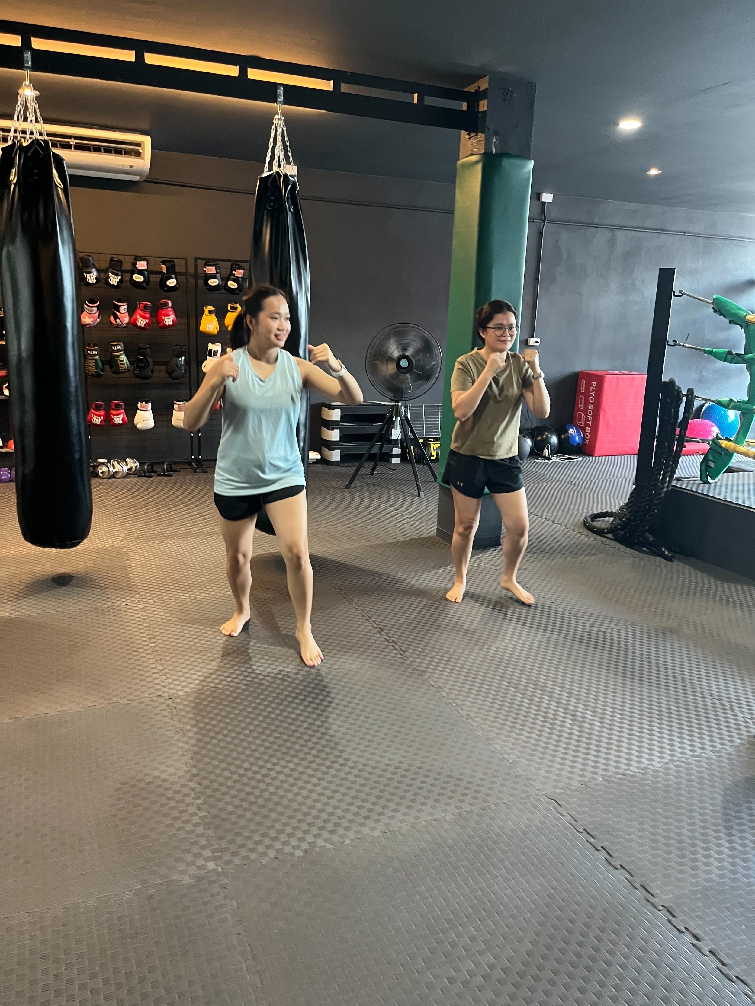 Two women in athletic wear exercising with fists raised inside a gym, with punching bags, boxing gloves, and workout equipment in the background.