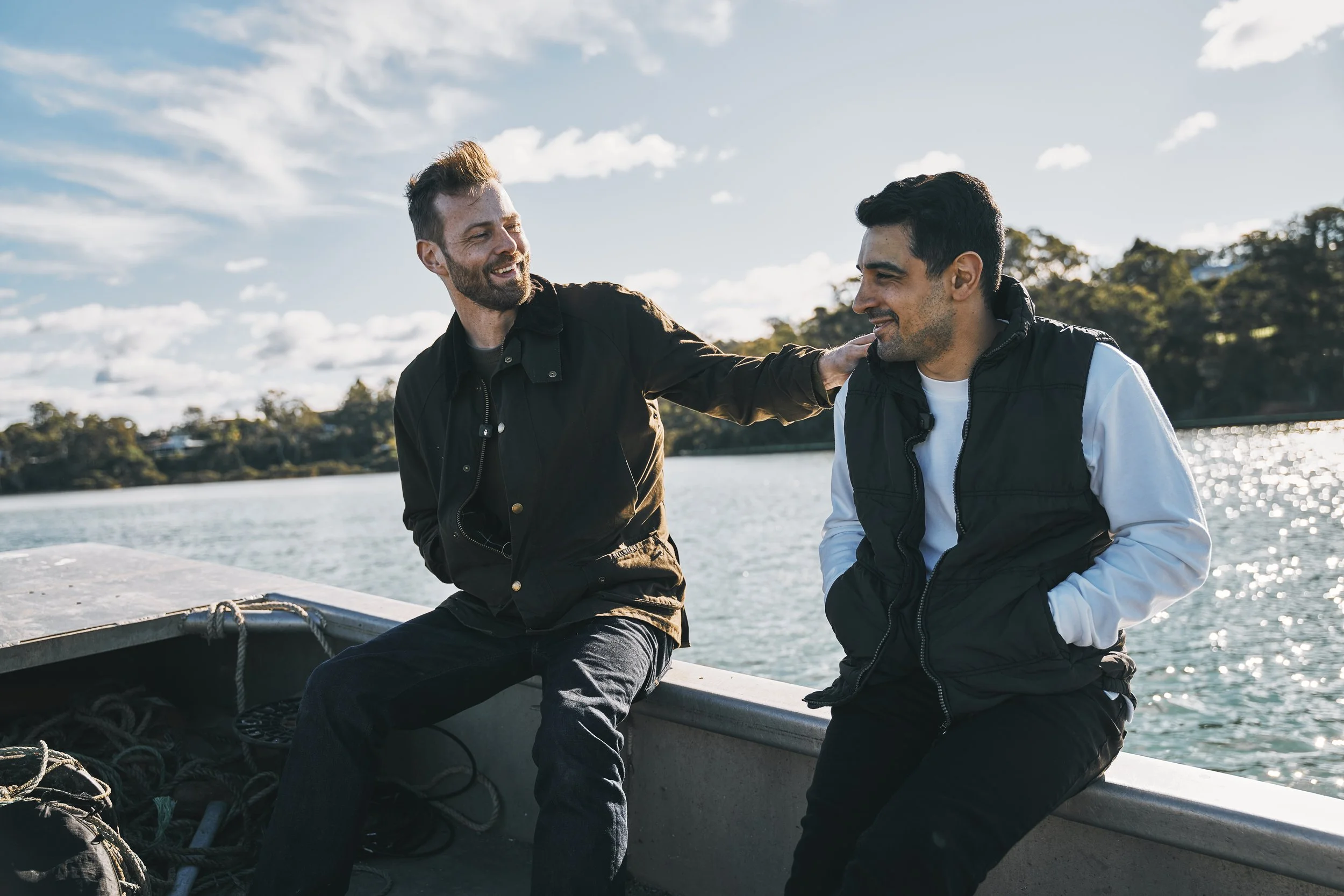 Two men sitting on a boat near a body of water, smiling and talking, with trees and a partly cloudy sky in the background.