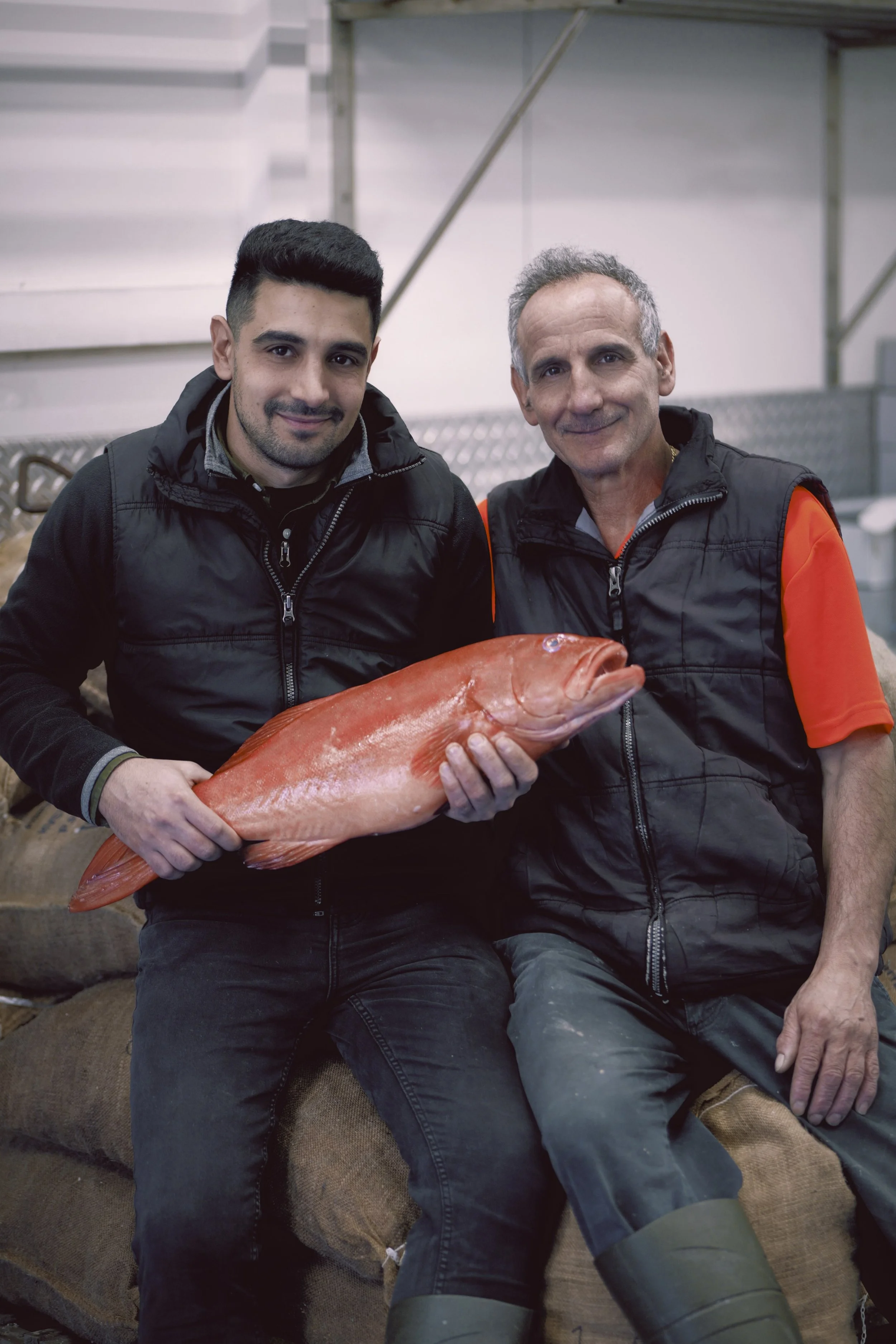 Two men sitting on a couch holding a large orange fish, with a background of stairs and a metallic structure.