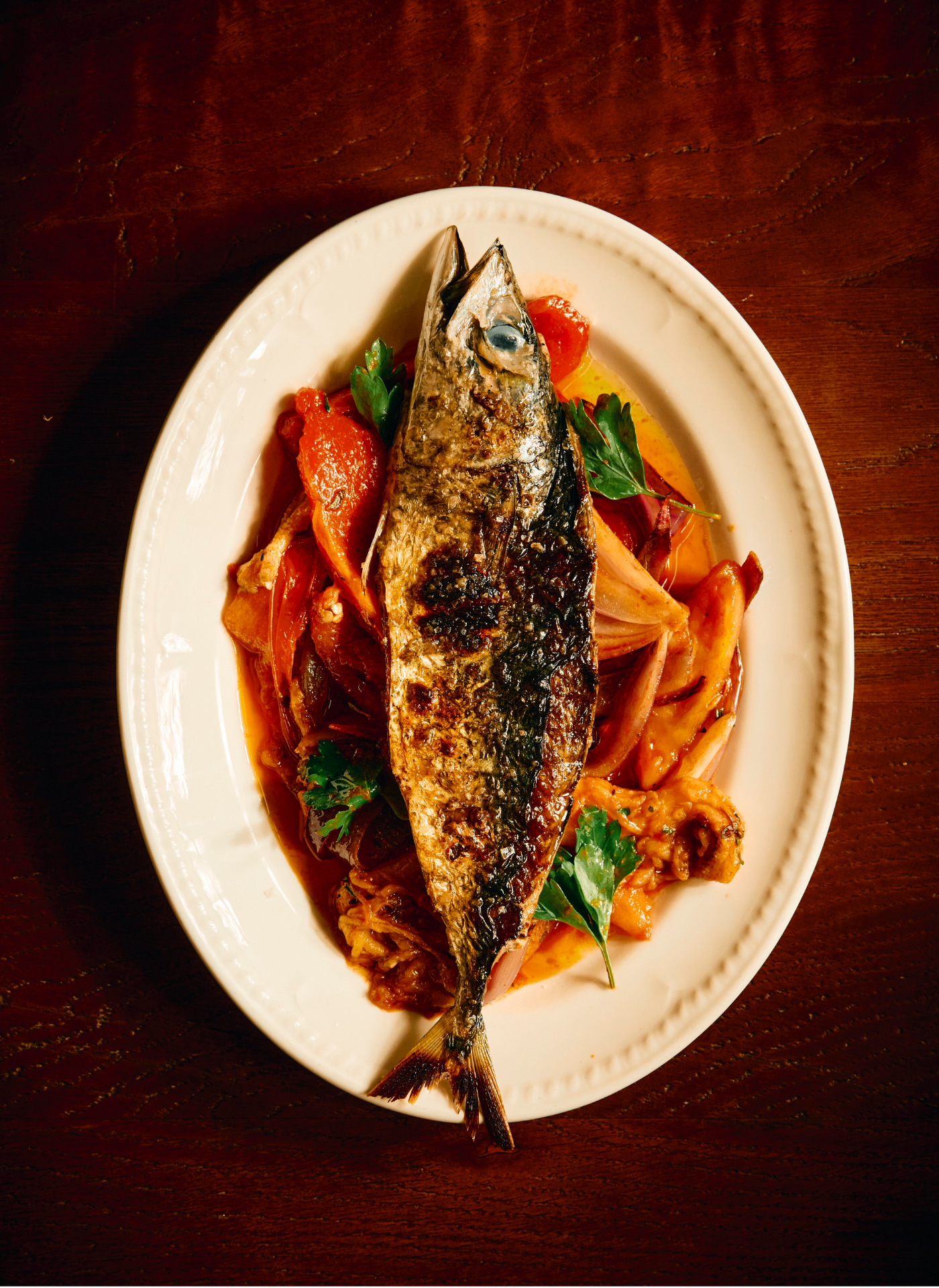 A whole grilled fish on a white oval plate, garnished with fresh herbs, served with vegetables in a sauce, on a dark wooden table.