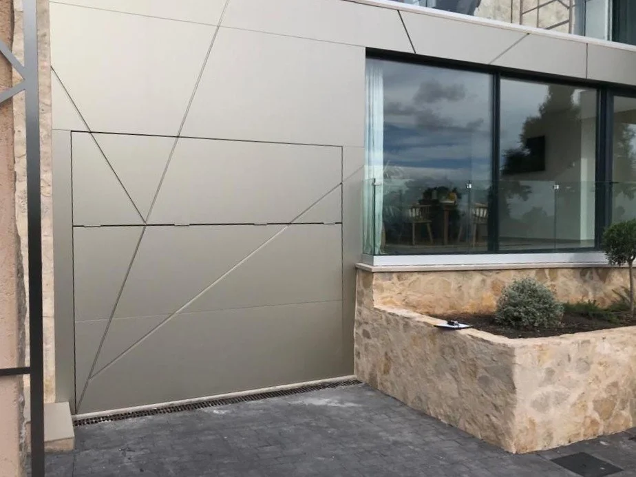 Fachada moderna con pared de vidrio y puerta metálica decorada con líneas diagonales, junto a un macizo de piedra con plantas ornamentales.