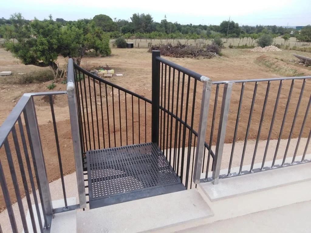 Barandal metálico en un puente o pasarela con vista a un campo con árboles y tierra árida en un área rural.