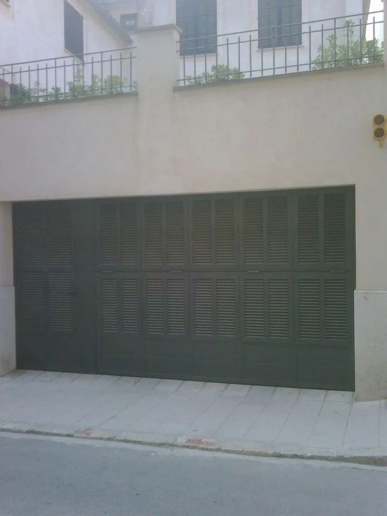 Puerta de garaje de color verde oscuro en un edificio residencial, con un muro de color claro y una reja en el balcón superior.