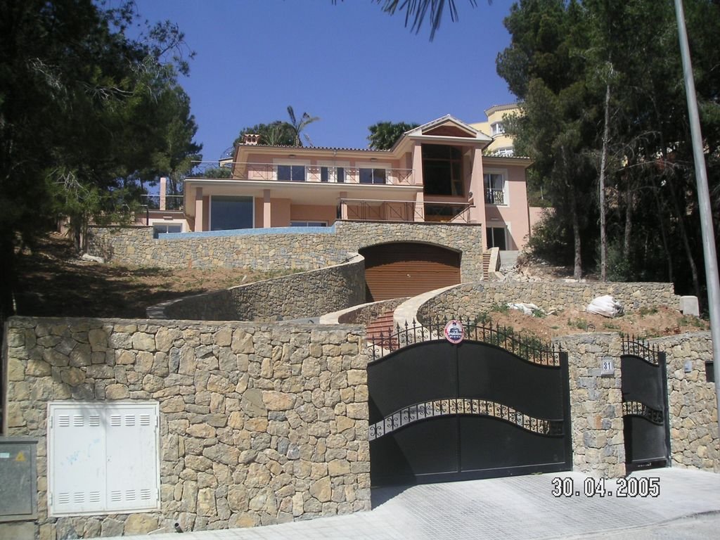 Casa moderna de varios pisos con fachada color rosa, puerta de garaje en arco, escalera en el lado derecho, y muro de piedra en el frente, rodeada de árboles.