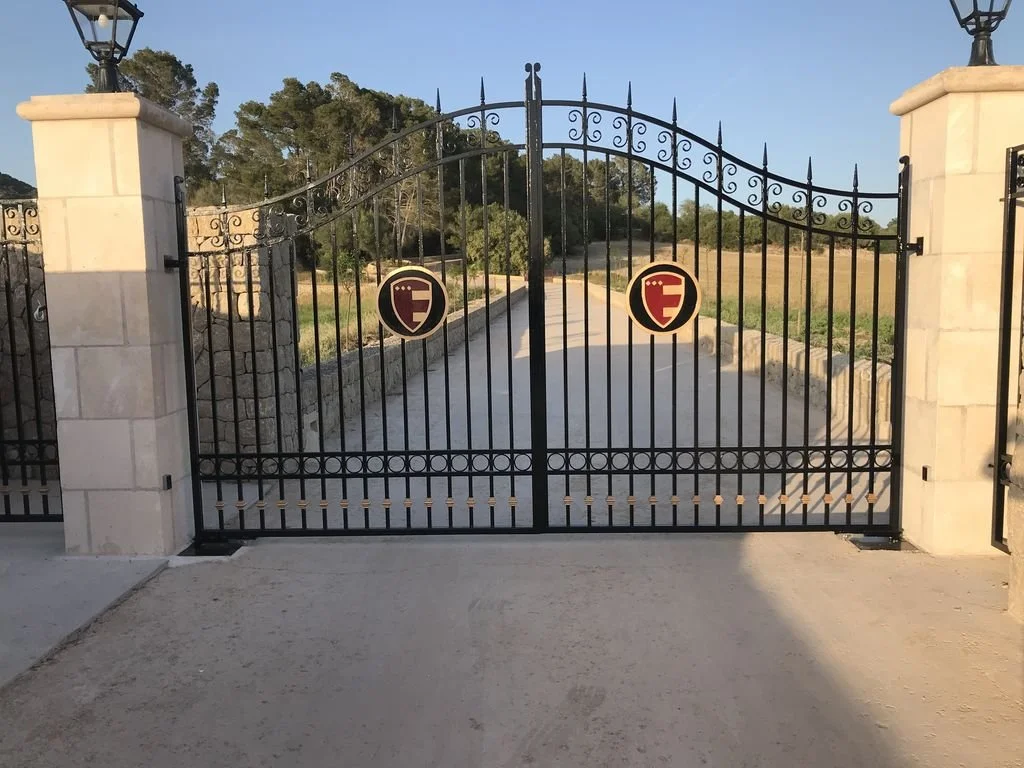 Puerta de rejas de metal con emblemas, paredes de piedra a los costados, camino de tierra y naturaleza al fondo.
