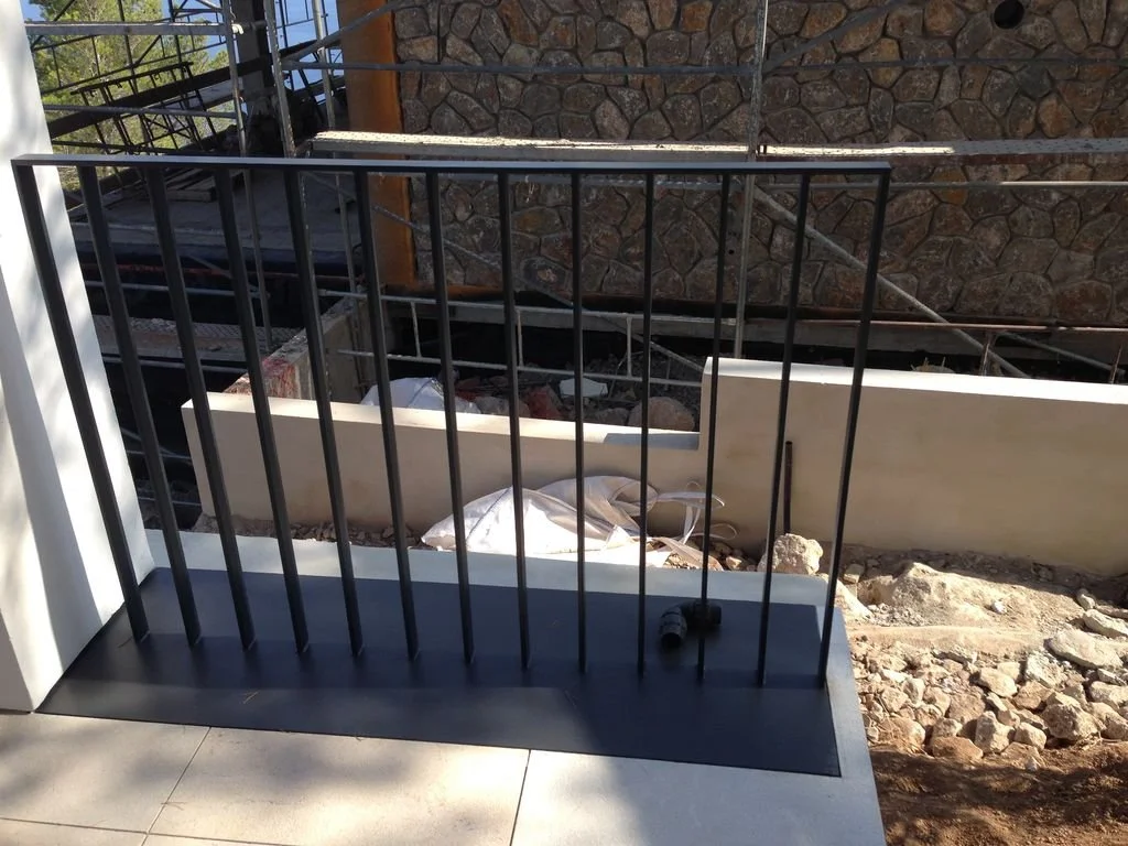 Barandal negro en una terraza, con construcción y piedras en el fondo.