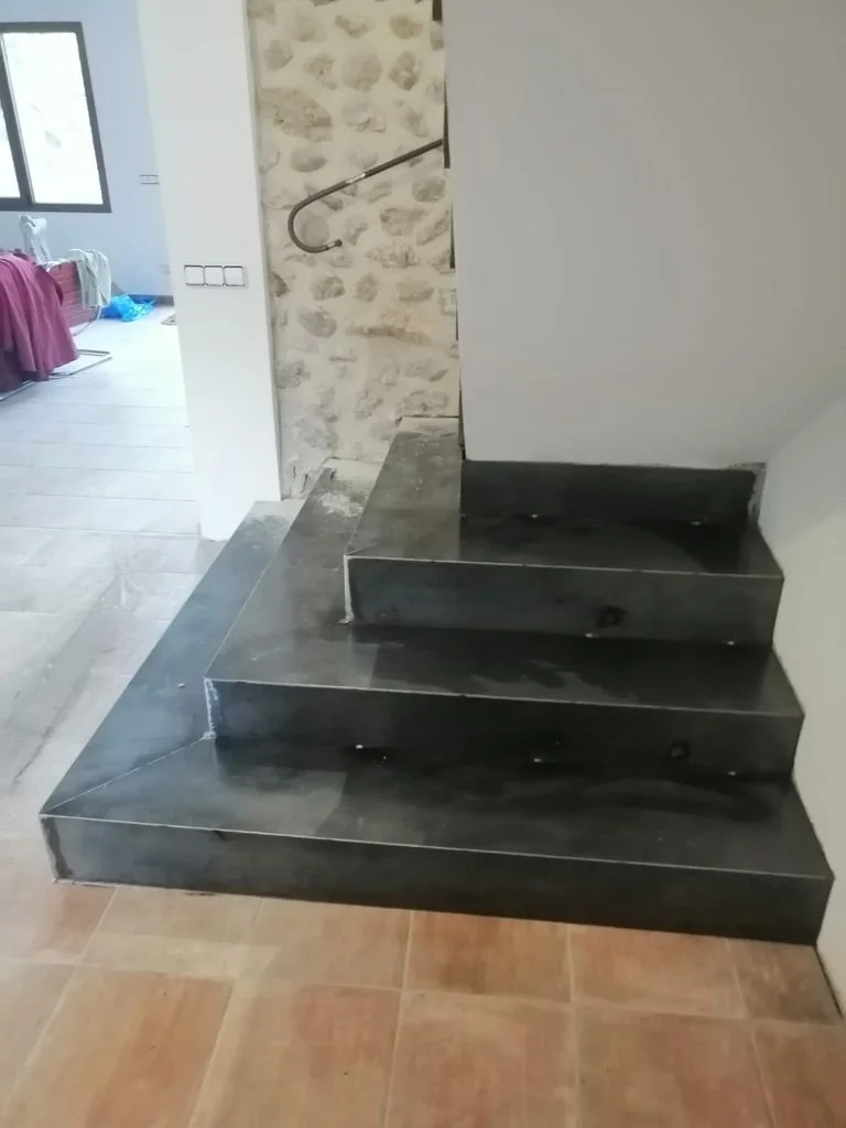 Escalera interna con pasos de piedra ennegrecida en una casa, con pared de ladrillos decorativos y piso de baldosas de terracota.