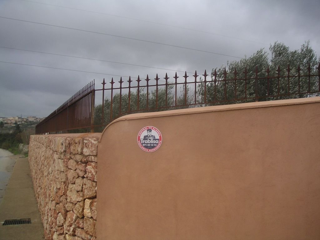 Valla de piedra y muro de color tierra con rejas de hierro y advertencia de seguridad y vigilancia.