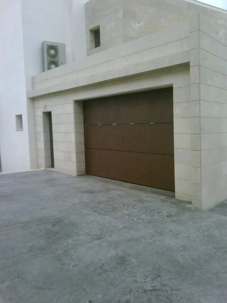 Fachada de una casa moderna con paredes de piedra beige y puerta de garaje de color marrón, con un suelo de cemento en el frente.