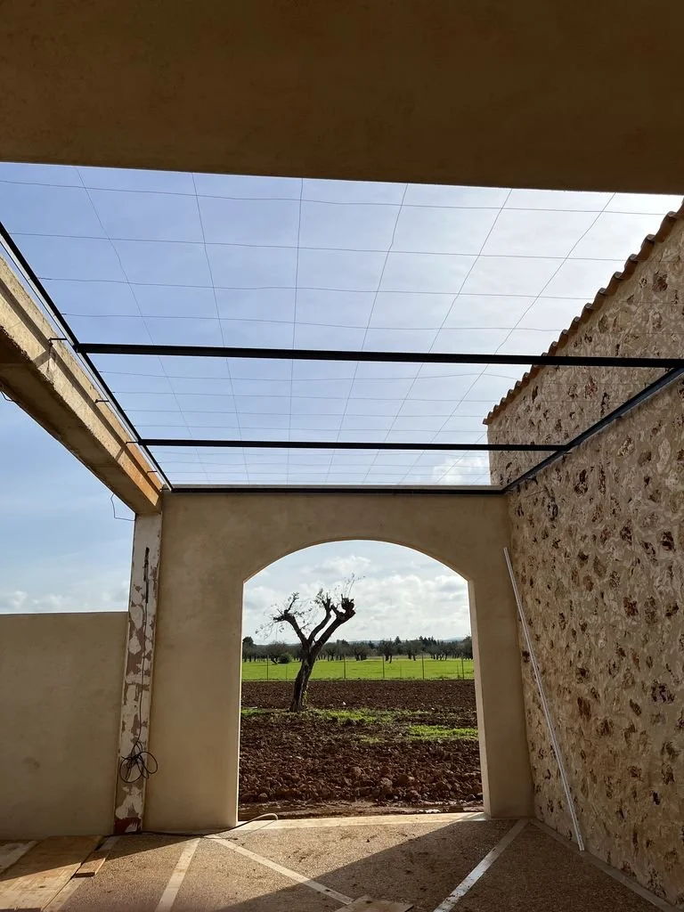 Vista de un patio interior en construcción con un arco abierto que revela un árbol solitario en un campo verde bajo cielo nublado.