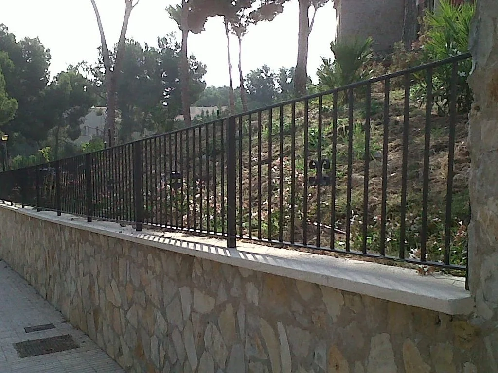 Reja de hierro sobre muro de piedra que delimita un área ajardinada con árboles y plantas.