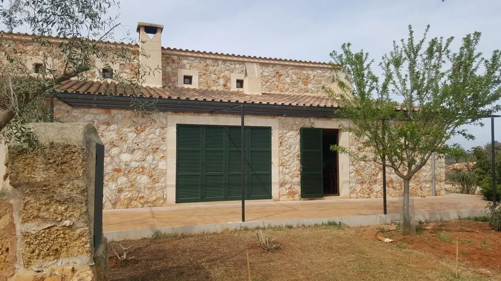 Casa de piedra con ventanas y puertas con celosías verdes, en un entorno rural con un árbol y un muro de piedra.