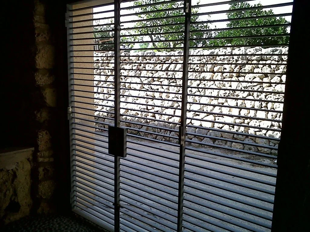 Puerta de metal con rejas y vista de una pared de piedras y árboles en el exterior.