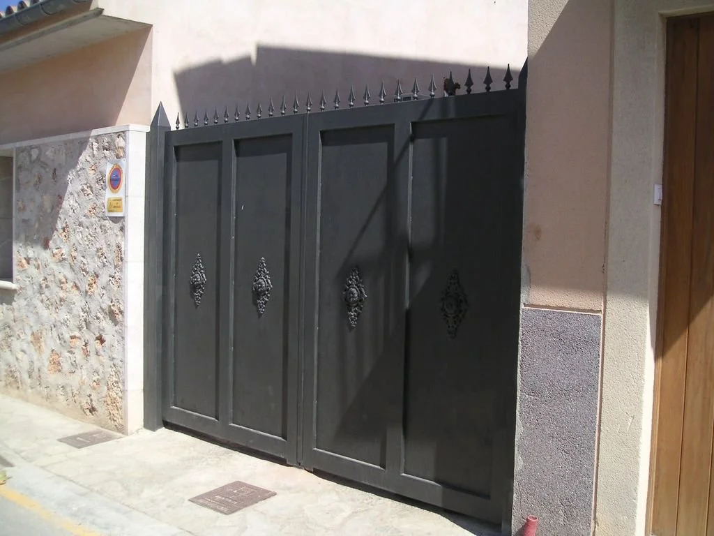 Puerta de metal negra con picos en la parte superior y decoraciones en el centro, en una entrada de casa en una calle.