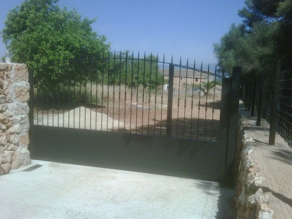 Valla de hierro negra con puerta, rodeada de muro de piedra y un camino de cemento, con árboles y cielo despejado en el fondo.