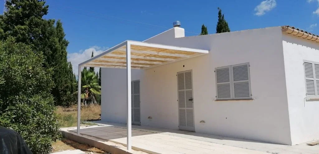 Casa blanca de una planta con ventanas con persianas grises y una puerta de metal, con una estructura de sombra en el patio y árboles verdes al fondo.