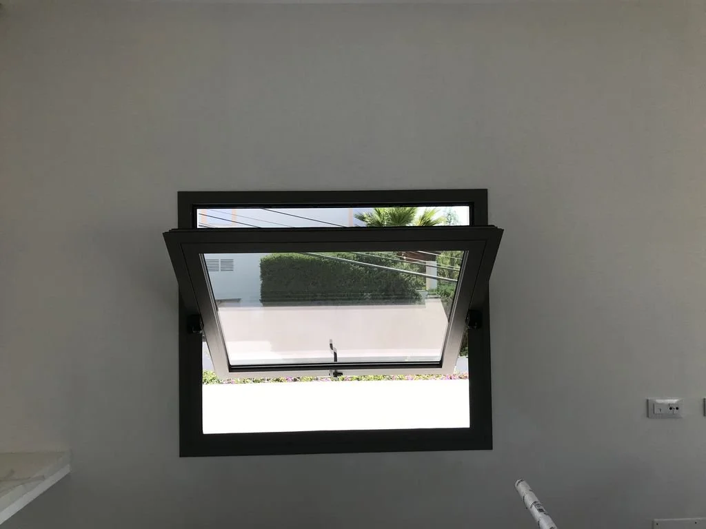 Ventana oscilante abierta en una pared blanca que muestra un jardín exterior con arbustos y palmera