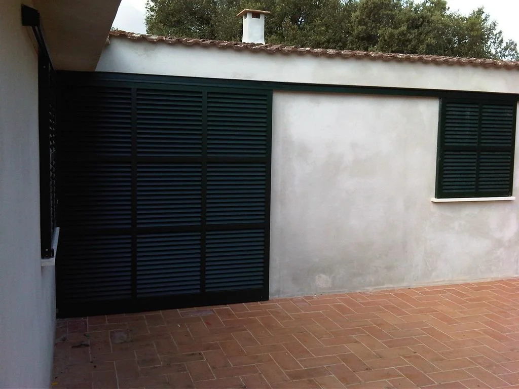 Pared exterior de una casa con ventanas con persianas y un portón negro de lamas horizontales en un patio con piso de ladrillos.