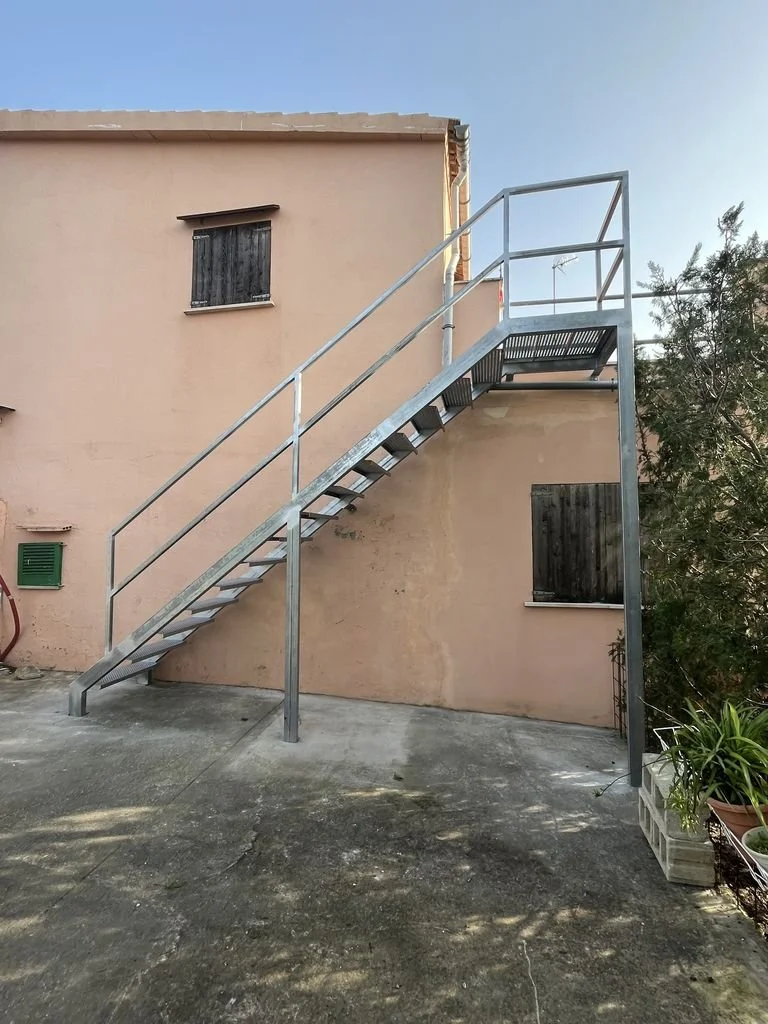 Escalera metálica exterior junto a pared de una casa con ventanas con contraventanas de madera.