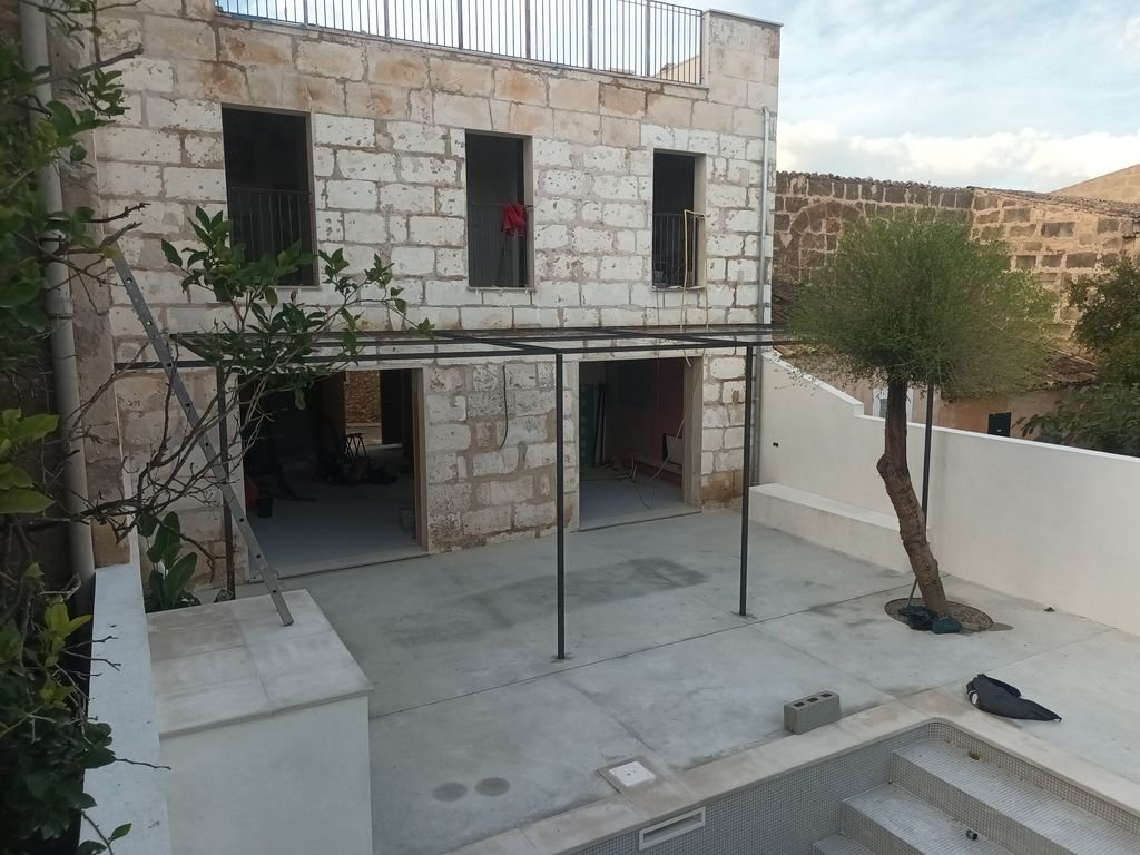 Patio con estructura metálica para baño o sombra, árbol en el rincón y casa en construcción con ventanas abiertas.