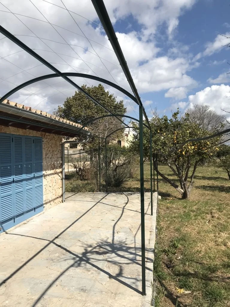 Estructura de malla de sombra en un patio con sombra de árboles y casa con paredes de piedra y puertas azules.