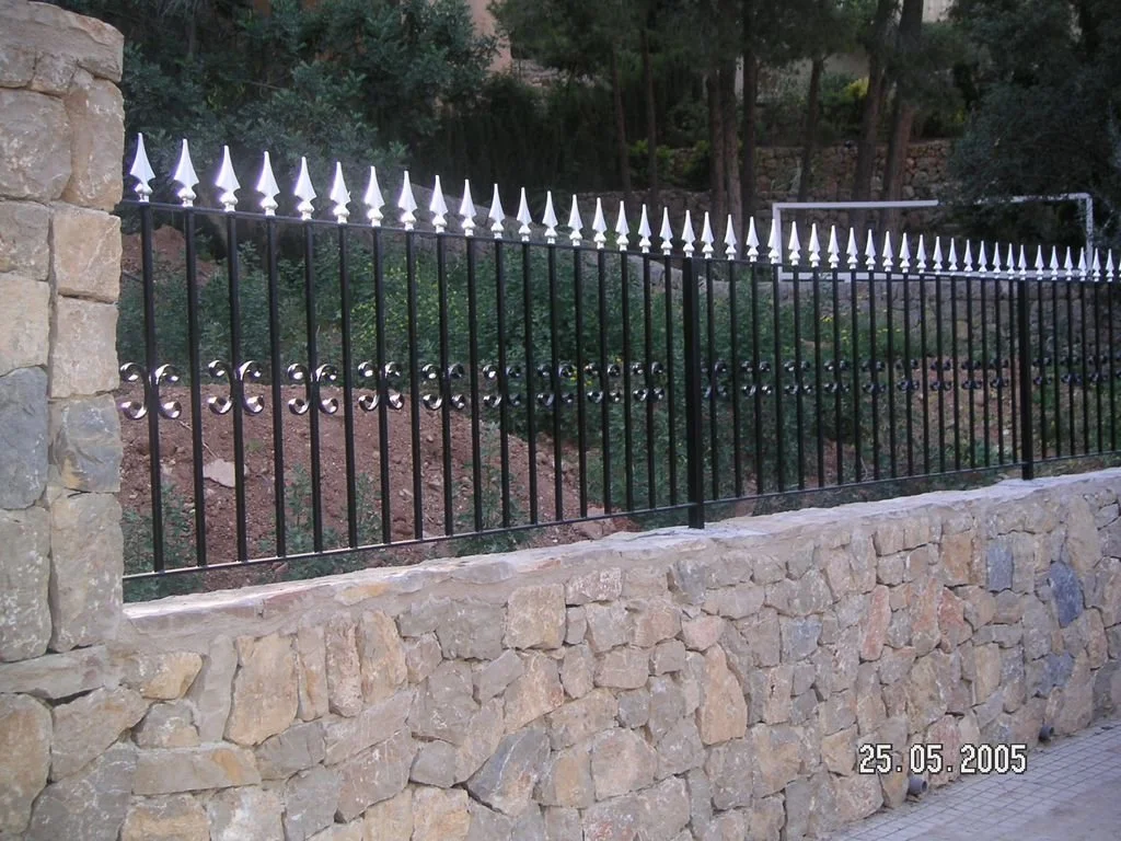 Una verja negra con pinchos blancos decorativos en la parte superior, instalada sobre un muro de piedras, con árboles y vegetación en el fondo.
