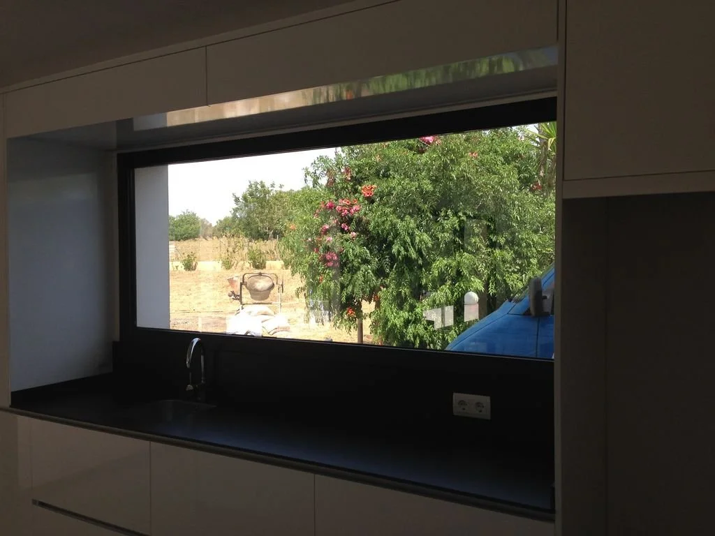 Vista desde una ventana en una cocina, con árboles y plantas verdes y flores rosadas afuera, incluyendo un árbol con flores y un vehículo azul parcialmente visible.