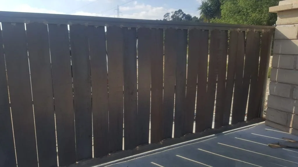 Una cerca de madera pintada de negro en un patio exterior, con algunas tablas inclinadas y un cielo parcialmente nublado.