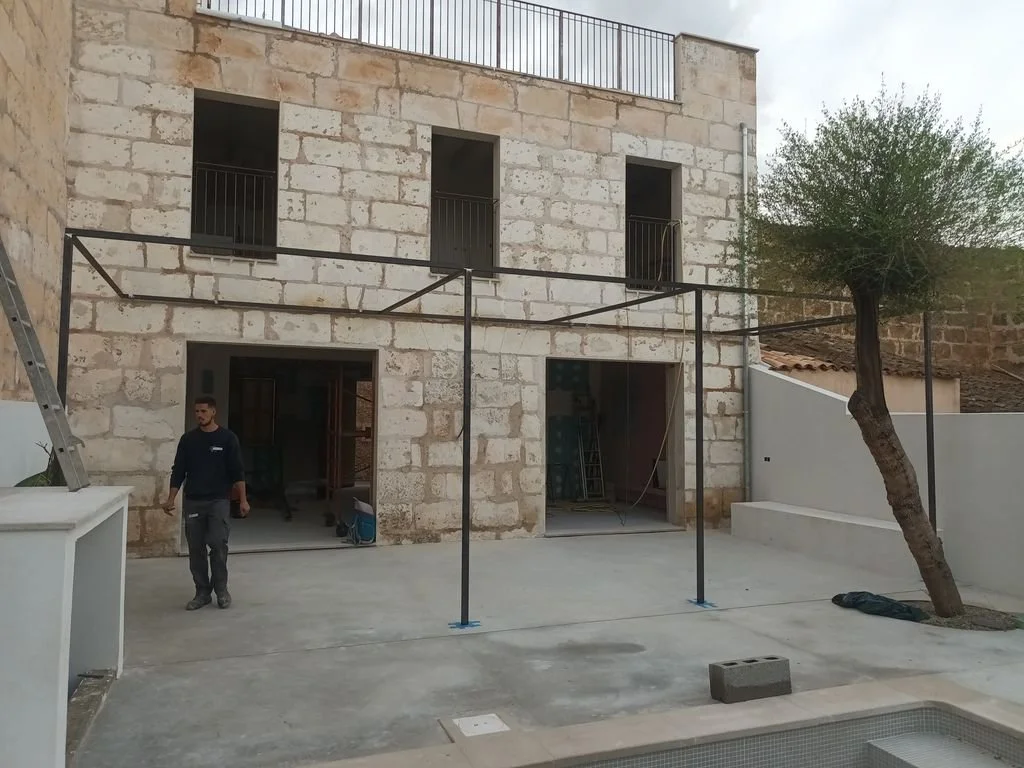 Construcción o remodelación de una casa con fachada de piedra y estructura metálica en el patio trasero, con un trabajador en el sitio.