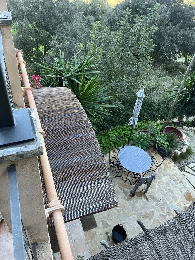 Vista desde arriba de un patio con mesa y sillas negras, paraguas cerrado, plantas y árboles verdes.