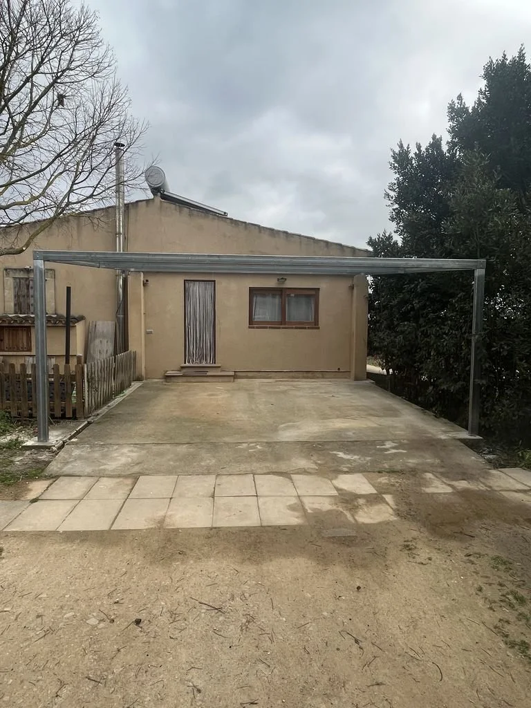 Estacionamiento con estructura de metal para automóvil, área de concreto y ladrillos en la entrada frente a una casa sencilla, en un día nublado.