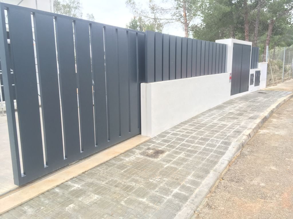 Muro de metal pintado de gris con una puerta de acceso, junto a una acera y un camino con pavimento de bloques, en un área con árboles y un fondo de paisaje natural.