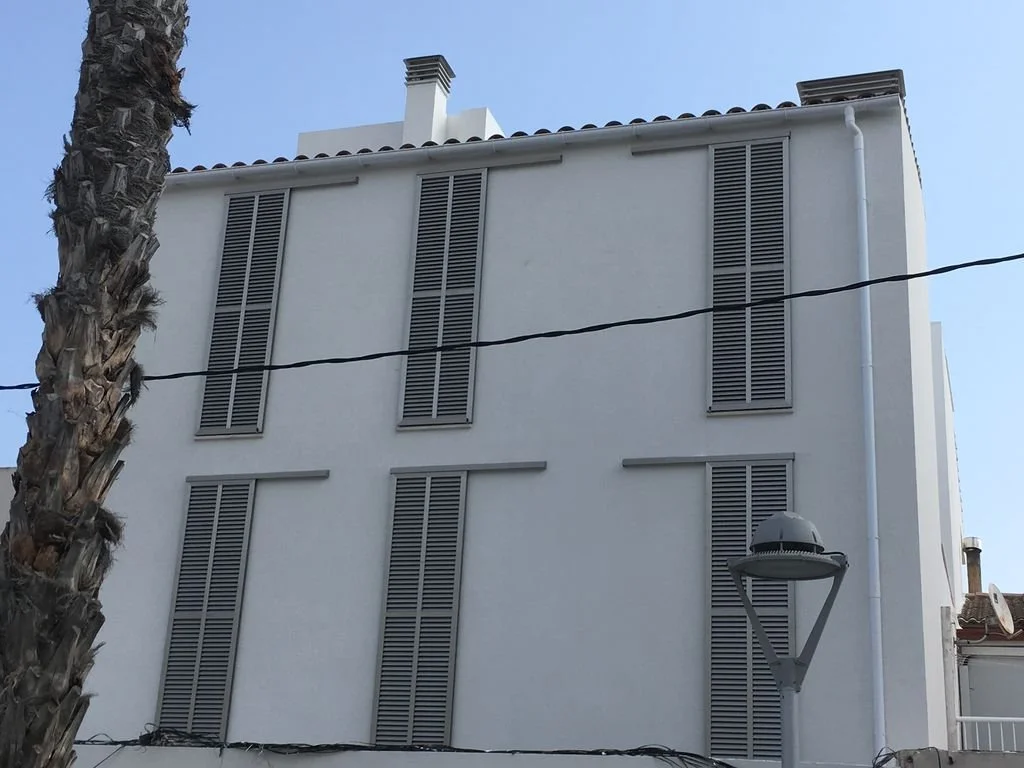 Edificio de color blanco con ventanas de persiana gris, una palmera a la izquierda, un poste de luz y cables eléctricos en la calle.
