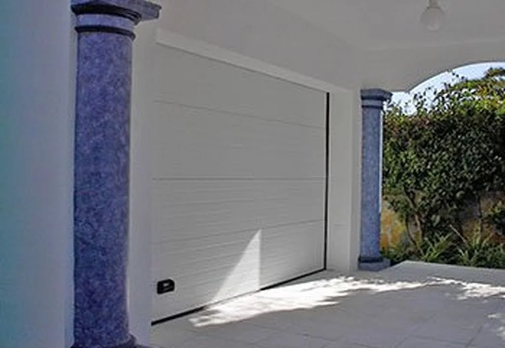 Puerta de garaje blanca en una casa con columnas decoradas y vegetación en el fondo.