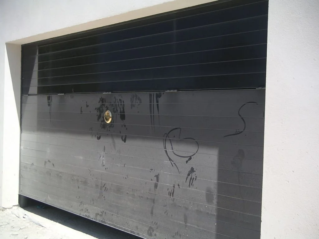 Puerta de garaje metálica en color negro con marcas de tinta y una mancha, instalada en una estructura de pared blanca.