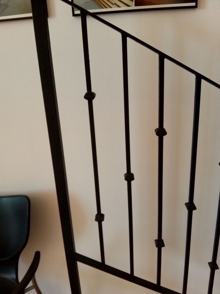 Barandal de reja negro en un interior, con pared blanca y una silla en la esquina izquierda.